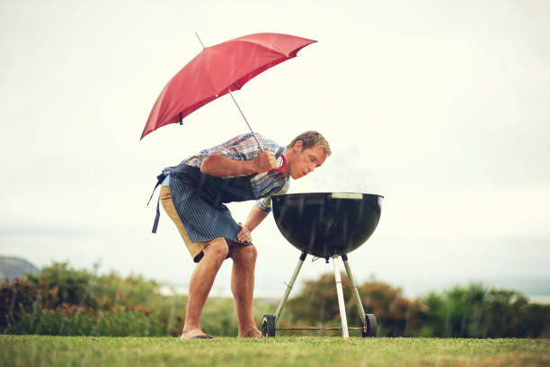 Who said braaing is always easy? Shot of a man having a barbecue outside in the rain while holding an umbrella and blowing on the fire south african braai stock pictures, royalty-free photos & images