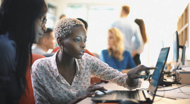 Graphic designers at work. Closeup side view of two mid 20's  black women doing their design project on a computer. They might be software developers as well.  Blurry coworkers in background, all released. digital transformation stock pictures, royalty-free photos & images