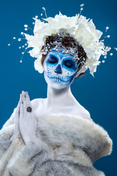 Santa Muerte woman at blue background Santa Muerte concept of body art. Young woman with short curly hair and creative visage. White flowers in hair, make up, portrait at blue background, wearing fur coat. muerte stock pictures, royalty-free photos & images