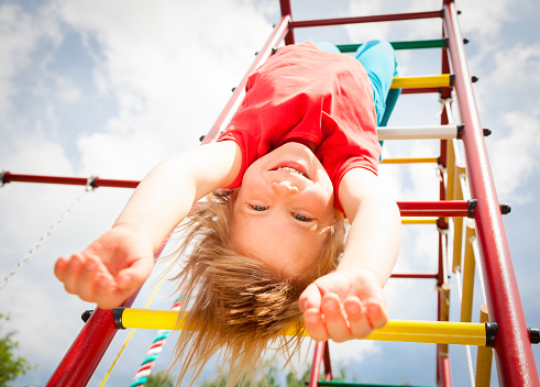 Low angle view of happy girl hanging from a climbing frame in a playground looking at camera smiling enjoying summertime