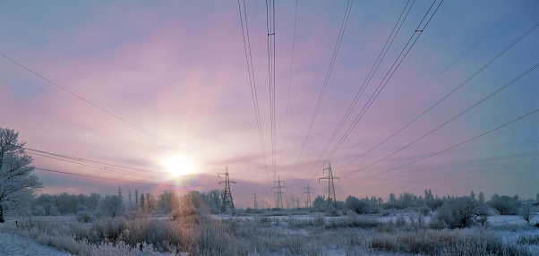 long lines of powerline towers stretching across a Winter Landscape. Sun shines through clouds.