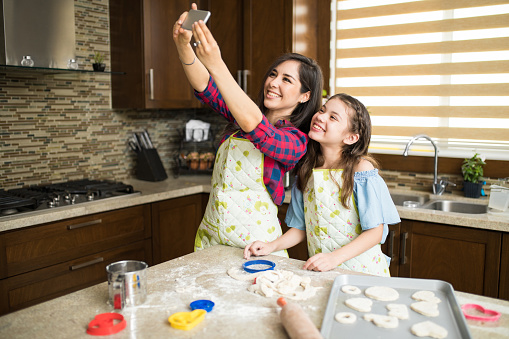 Pretty single mother and her daughter baking together at home and taking a selfie with a smartphone