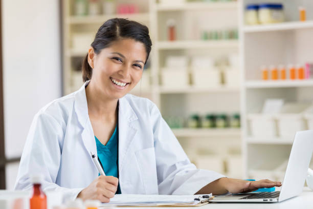 Female pharmacist uses laptop to check inventory Pretty mid adult female Asian pharmacist uses a laptop at work. pharmacy tech stock pictures, royalty-free photos & images