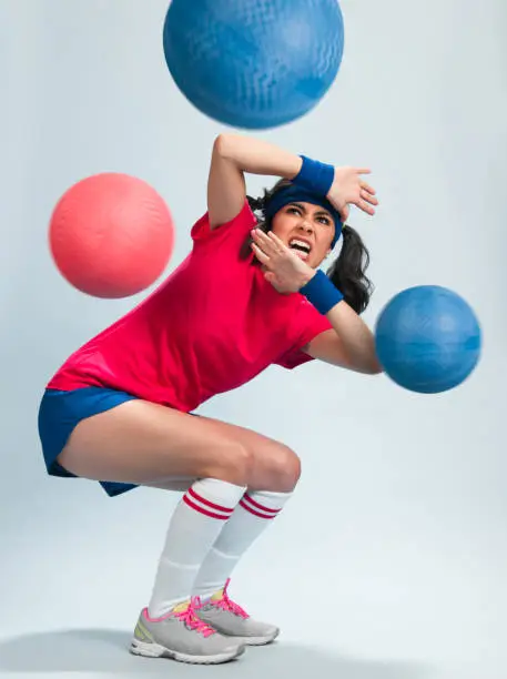 Young Hispanic Women Playing Dodgeball in a retro gym outfit with multipe balls thrown at her.