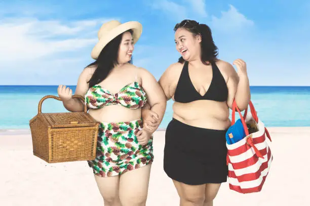 Two fat women walking on sand beach while holding picnic basket and beach item on bag
