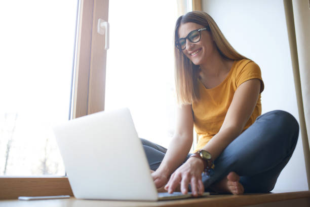 istock Smiling young woman sitting on window sill with computer 694319630
