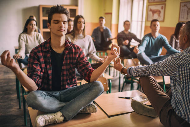 Relaxed high school students exercising Yoga in the classroom. Large group of students sitting in Lotus position with their teacher and exercising Yoga. teen yoga stock pictures, royalty-free photos & images