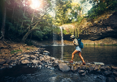 Shot of a young woman walking on a rocky surface towards a waterfall in the forest