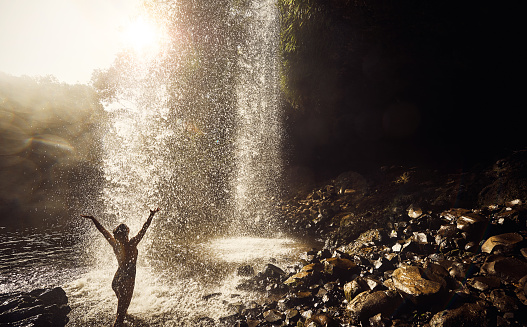 Rearview shot of a young woman arms raised underneath a waterfall enjoying a day in nature