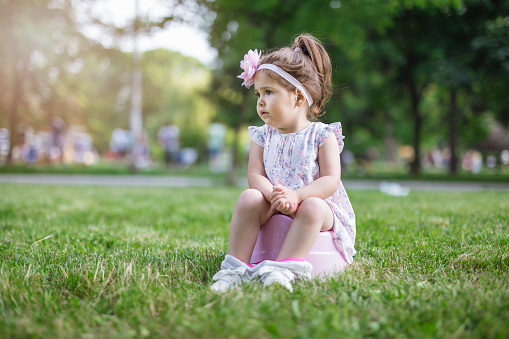 24 months old cute toddler girl is stopping to wear diapers, so potty is must have everywhere. She is urinating in a public park on a warm spring day.The emotion is real and candid because so is used to being photographed