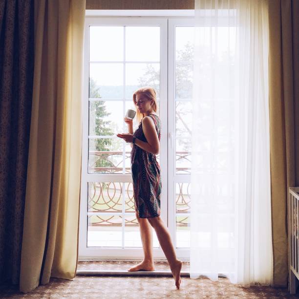 istock Young blonde woman standing by the window at home. 694426310