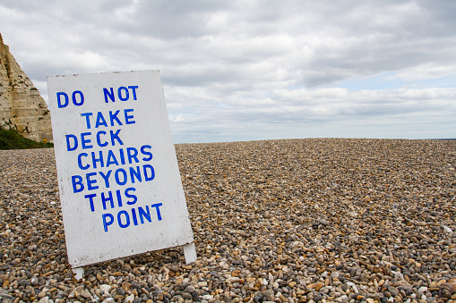 A worn and weathered hand made sign on a deserted pebble beach forbidding people from taking their deckchairs beyond a certain point into a dangerous area.