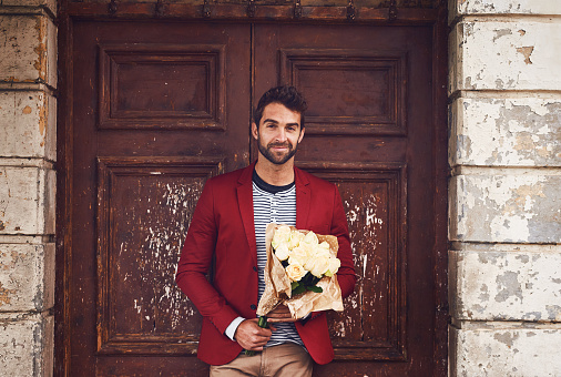 Shot of a handsome young man standing outside a door with a bunch of flowers looking smart and happy