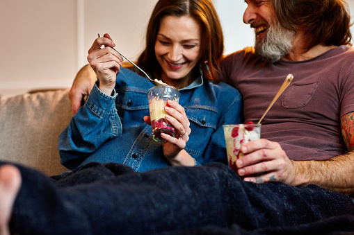Happy mature couple sitting together on sofa and eating ice cream. Pregnant woman with husband relaxing in living room and having ice cream.