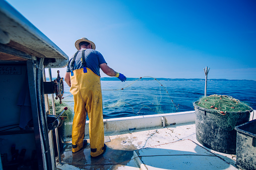 Fisherman pulling the net out of water. He is standing on his boat. Sea and blue sky in back.