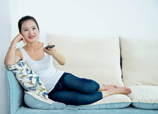 istock Young asian woman with remote control watching television on sofa 694478336