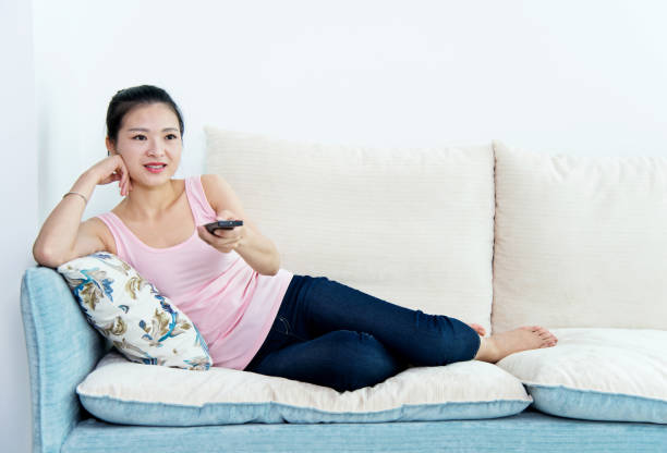 istock Young asian woman with remote control watching television on sofa 694481720