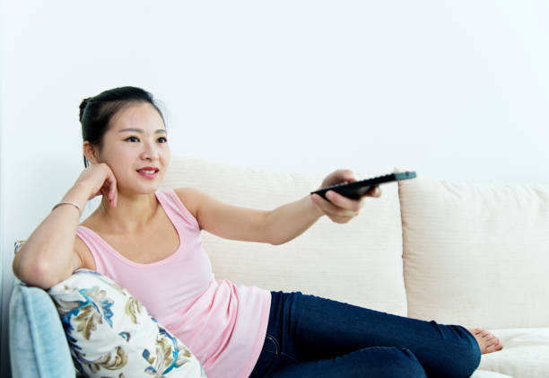 istock Young asian woman with remote control watching television on sofa 694481884
