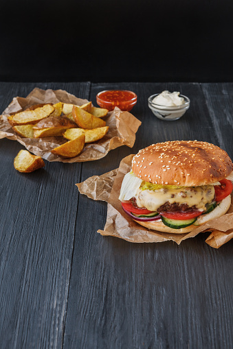 Fast food restaurant dish top view. Meat cheese burger in craft paper, potato chips and wedges. Take away set on dark black wood background. Hamburger and spicy tomato sauce. Vertical
