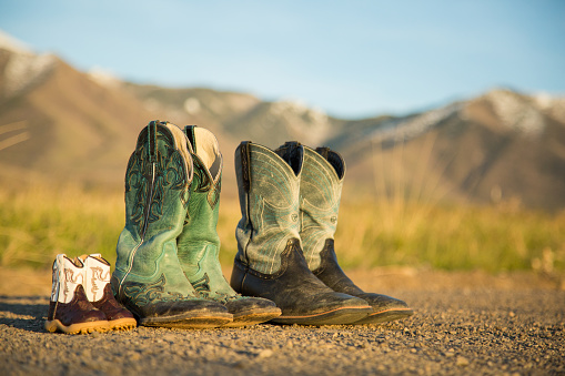 Three pair of cowboy boots on a dirt road, with long shadows from late afternoon sun and mountains in background. One pair of baby boots, one pair of female boots that are turquoise and brown, and one pair of male boots that are a lighter turquoise and black. Suggests a family who live a western lifestyle.