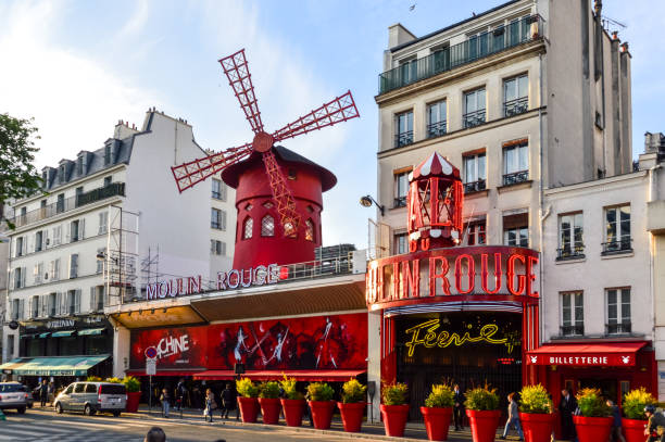 The Moulin Rouge during a sunny day. Moulin Rouge is a famous cabaret built in the 19th century, and is located in the Paris red-light district of Pigalle. Photo contains some people and vehicles.