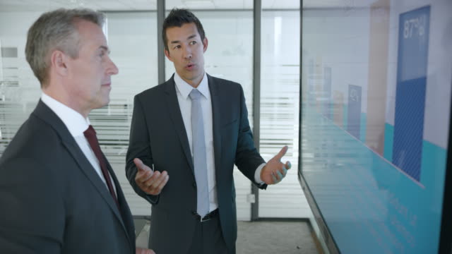 Senior Caucasian man and Asian male colleague discussing the numbers shown in the financial presentation on the large screen in the meeting room