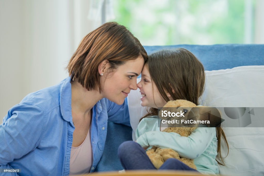 Mother And Daughter A young Caucasian girl is indoors in a hospital room. She is wearing casual clothing. She is lying in bed and holding a stuffed dog. She is rubbing noses with her mother. Child Stock Photo