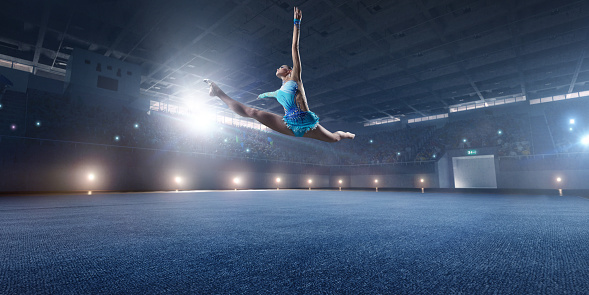 A gymnast girl makes a leap on a large professional stage. She is wearing a professional swimsuit for gymnastics. Behind her is a stadium with spectators.