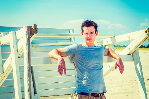 Man waiting for you. Wearing a gray t shirt, arms resting on a wooden stick, a young handsome guy is standing by a wooden structure on the beach, narrowing eyes, charmingly looking at you.