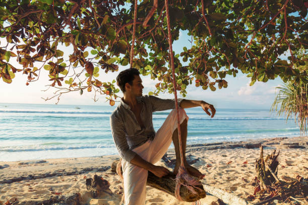 istock Attractive handsome guy sitting on the swing on the tropical beach. 694742542