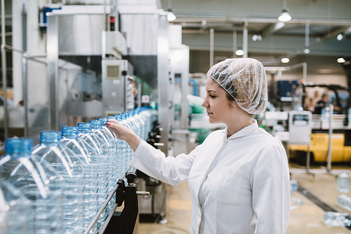 Young happy woman worker checking robotic line for bottling and packaging pure drinking water into bottles and canisters.