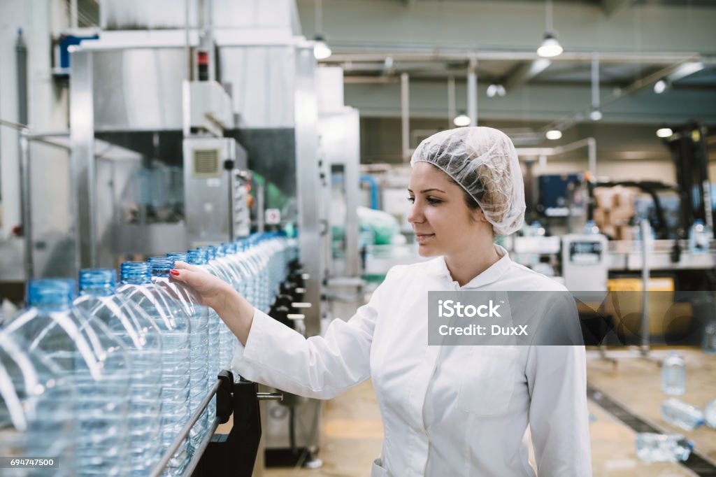 Young woman factory worker Young happy woman worker checking robotic line for bottling and packaging pure drinking water into bottles and canisters. Bottling Plant Stock Photo