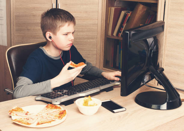 gosse manger pizza et surfer sur internet ou watshing vidéo sur ps. boy dans les écouteurs manger restauration rapide tout en utilisant l’ordinateur dans sa chambre. - computer chip photos et images de collection