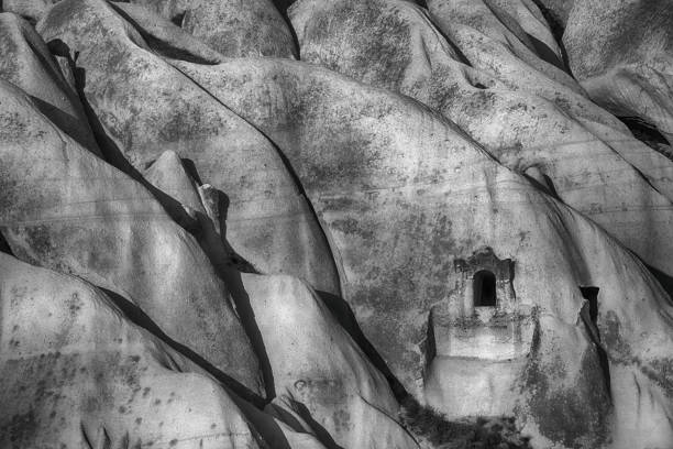A unique rock formation stands prominently, featuring a cave dwelling carved into its side. Surrounding greenery and dry grasses hint at the natural beauty of Cappadocia under cloudy skies.