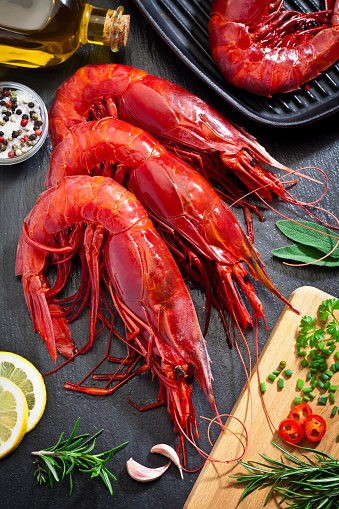 Top view of three giant red shrimp shot on dark table. An iron grill with one shrimp is visible at the upper-right corner of the frame and a wooden cutting board with herbs and spices is at the bottom-right corner. Some ingredients for cooking shrimps like olive oil, salt and pepper, garlic, lemon and some herbs are all around the shrimps. DSRL studio photo taken with Canon EOS 5D Mk II and Canon EF 100mm f/2.8L Macro IS USM