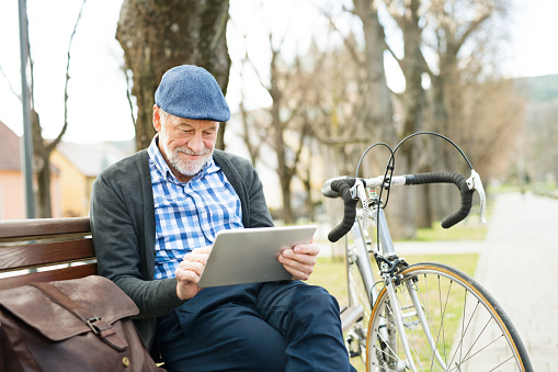 Handsome senior man with bicycle in town park sitting on bench, working on tablet. Sunny spring day.