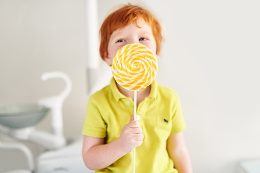 Cute candy lover hiding behind big yellow lollipop in dentists office