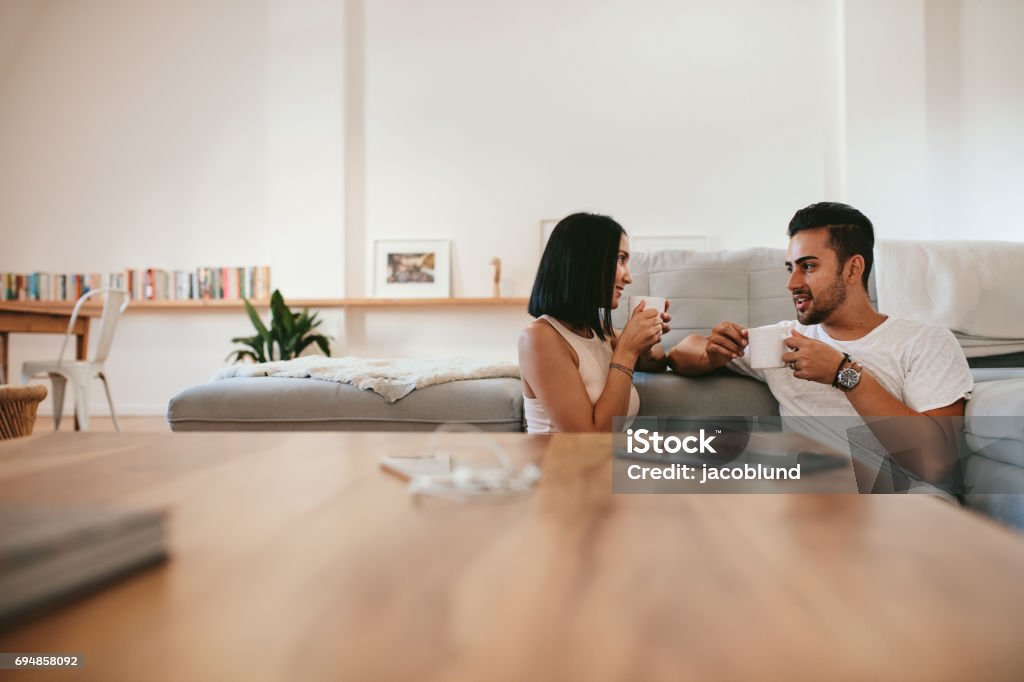 Young couple having coffee at home Young couple relaxing by the couch with cup of coffee in the living room. Man and woman sitting together at home having coffee and talking. Young Couple Stock Photo