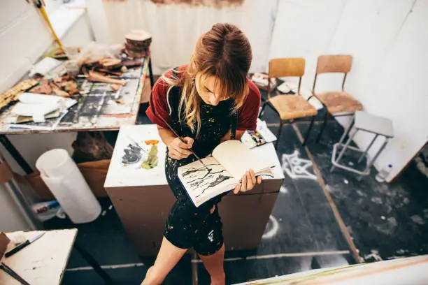 Female artist drawing pictures in her workshop. Young woman sitting and painting in her drawing book.