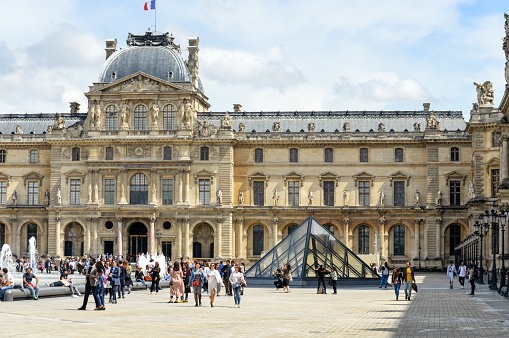 The Louvre is one of the most famous monuments and tourist attractions in Paris. This photo was taken during a warm spring overcast day in the museum courtyard and features the grandoise architecture of the palace. It also shows the many visitors that this place receives every day.