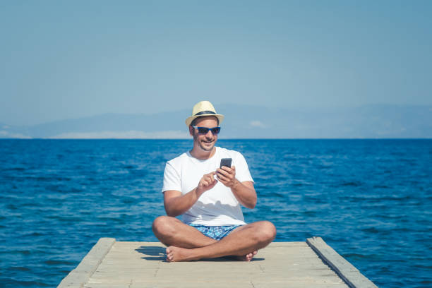 istock Tourist guy sitting on dock and using smartphone. Summertime and technology concepts. 694933876