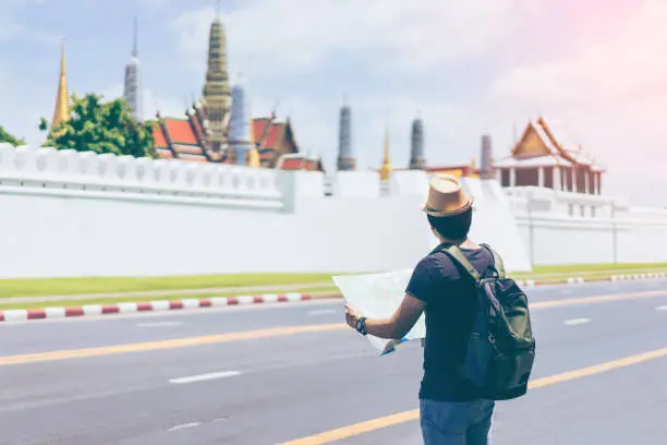 Photo of Young man traveler with backpack and hat looking the map with Grand palace and Wat phra keaw at Bangkok Thailand