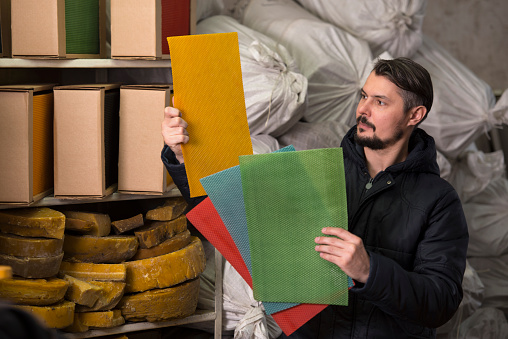 Factory man holding and examining color beeswax candle sheets.