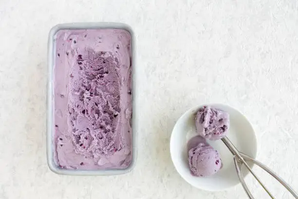Homemade Blueberry Ice Cream in metal container and two scoops in a white porcelain bowl on white marbled background.