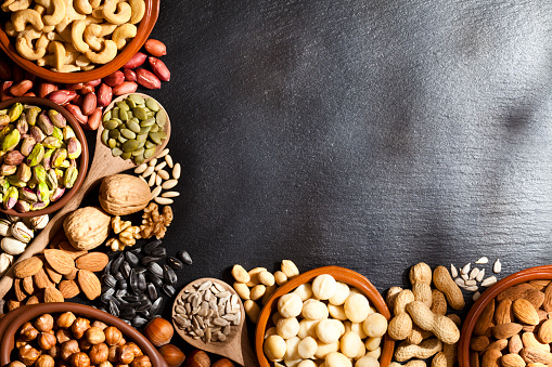 Horizontal shot of a black slate table with a large assortment of nuts arranged at the left and bottom borders in brown bowls, wooden spoon or placed directly on the background leaving a useful copy space at the center-right of the frame ready for text and/or logo. Nuts included in the composition are pistachios, hazelnut, pine nut, almonds, pumpkin seeds, macadamia nuts, sunflower seeds, peanuts, cashew and walnuts. Predominant color is brown and gray. DSRL studio photo taken with Canon EOS 5D Mk II and Canon EF 100mm f/2.8L Macro IS USM