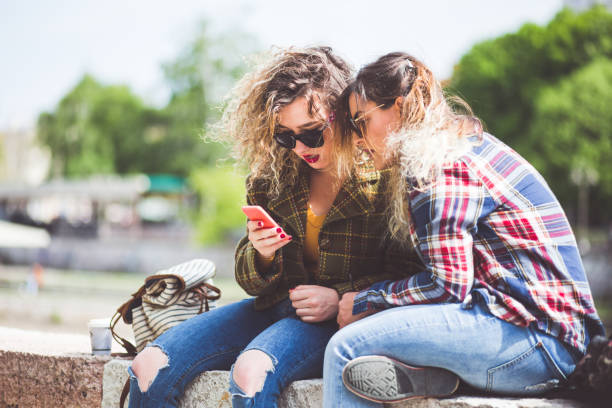 istock Girfriends Surfing The Net Using Phone While Sitting On River Quay 695078930