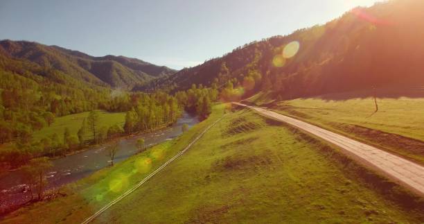 meados voo de ar fresco da montanha rio e prado na manhã ensolarada de verão. estrada de terra rural abaixo - aerial view mid air farm field - fotografias e filmes do acervo