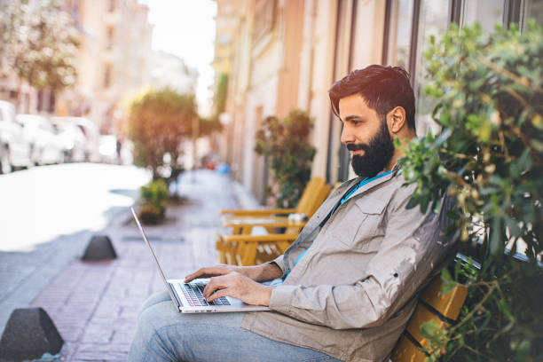 Portrait of young man with long beard working at cafe Portrait of a long beard middle eastern ethnicity hipster young man texting with smartphone and working laptop at outdoor urban coffee shop turkey middle east stock pictures, royalty-free photos & images