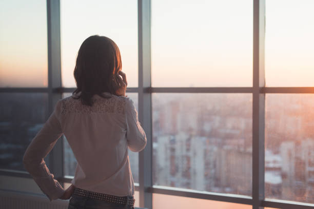 Rear view portrait of young worker speaking using cell phone, looking out the window. Female having business call, busy at her workplace in evening. Rear view portrait of young worker speaking using cell phone, looking out the window. Female having business call, busy at her workplace in evening office window night stock pictures, royalty-free photos & images