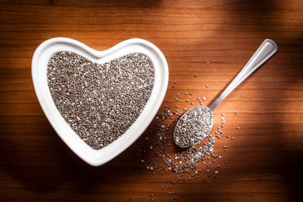 Top view of a heart shaped bowl filled with chia seeds placed at the left of a wooden table. A spoon with seeds is at the right. Predominant color is brown. Low key DSRL studio photo taken with Canon EOS 5D Mk II and Canon EF 100mm f/2.8L Macro IS USM